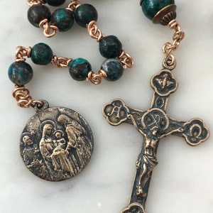 Puede incluir: Un rosario de tono cobre con cuentas de piedra azul y verde y un crucifijo de bronce con una representación de Jesucristo. El colgante central presenta una imagen de bronce de la Sagrada Familia.
