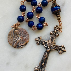 May include: A rosary with deep blue, spherical beads and copper-colored wire links. The rosary features a detailed bronze crucifix and a circular pendant with an image of a saint. The beads are approximately 0.3 inches in diameter.
