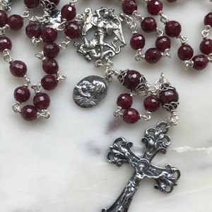 Puede incluir: Un rosario de plata con cuentas de piedras preciosas de color burdeos y un crucifijo de plata. El crucifijo presenta un diseño detallado y una figura de Jesucristo. El rosario también incluye un colgante de plata con una representación de un ángel.