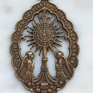 Puede incluir: Un colgante de bronce con un diseño recortado que presenta dos ángeles flanqueando una cruz con un sol radiante detrás. El colgante tiene el texto "CORTE DE AMOR Y EXPIACION AL SS. SACRAMENTO" alrededor del borde.