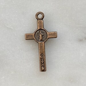 May include: A small, bronze-coloured cross pendant with a circular image of Saint Benedict in the centre. The cross has a loop at the top for attaching to a chain or cord.