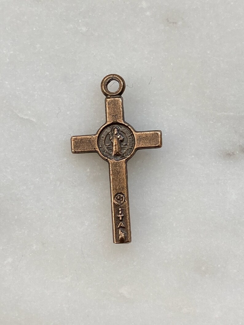 May include: A small, bronze-coloured cross pendant with a circular image of Saint Benedict in the centre. The cross has a loop at the top for attaching to a chain or cord.