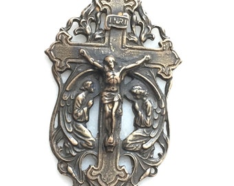 May include: A bronze pendant featuring a crucifix with a figure of Jesus Christ on the cross. Two angels are kneeling on either side of the cross, with their wings spread out. The pendant is ornate and detailed, with a filigree design around the edges. The pendant is marked "INRI" at the top.