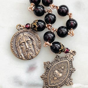 May include: A rosary featuring dark red, round beads strung on copper-colored wire. The rosary includes two antique-style bronze pendants, one with a portrait and the other with a heart design. The beads are approximately 8mm in diameter.