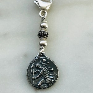 May include: Silver charm with a round, silver pendant featuring a detailed image of a saint. The charm is attached to a silver chain with silver beads and a silver clasp.