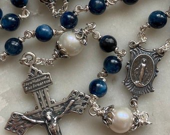 Puede incluir: Un rosario de plata con cuentas azules y negras y detalles de perlas blancas. El rosario presenta un crucifijo de plata con una figura de Jesucristo y una medalla de plata con una imagen de la Virgen María.