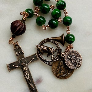 May include: A green stone rosary with a brown wooden bead, a bronze crucifix, and several bronze charms, including a horn, a saint medal, and a shepherd with a dog.