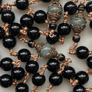 May include: A close-up of a necklace featuring large, glossy black beads strung together with copper-colored wire. The necklace also includes decorative, gray-green beads with ornate metalwork.