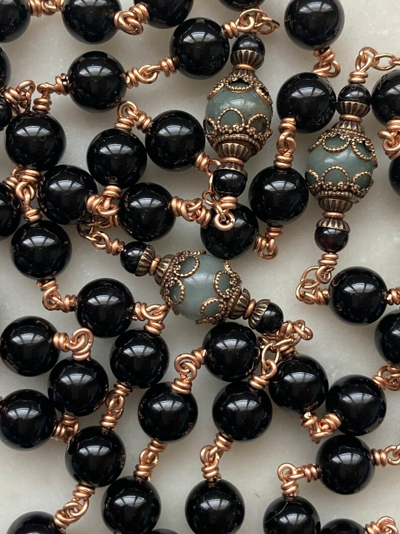 May include: A close-up of a necklace featuring large, glossy black beads strung together with copper-colored wire. The necklace also includes decorative, gray-green beads with ornate metalwork.
