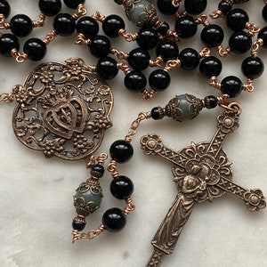 May include: A detailed rosary featuring black beads strung on copper wire. The rosary includes ornate bronze-colored metalwork, including a cross and a decorative centerpiece with a heart and crown design. Gray beads are interspersed throughout.