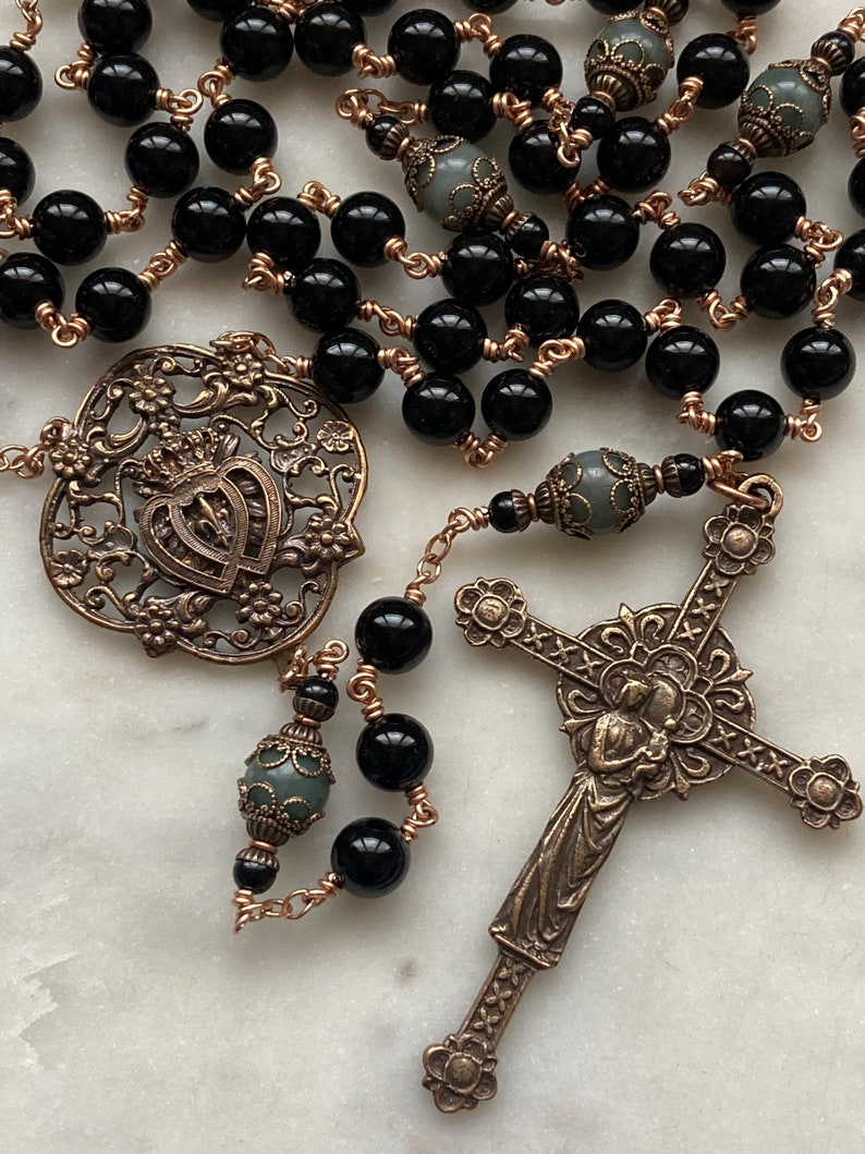 May include: A detailed rosary featuring black beads strung on copper wire. The rosary includes ornate bronze-colored metalwork, including a cross and a decorative centerpiece with a heart and crown design. Gray beads are interspersed throughout.