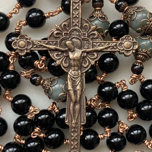 May include: A rosary featuring black, glossy beads strung on copper-colored wire. The centerpiece is a detailed bronze crucifix with the figure of Jesus. Additional gray beads with ornate caps are interspersed throughout.
