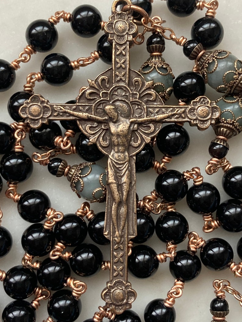 May include: A rosary featuring black, glossy beads strung on copper-colored wire. The centerpiece is a detailed bronze crucifix with the figure of Jesus. Additional gray beads with ornate caps are interspersed throughout.