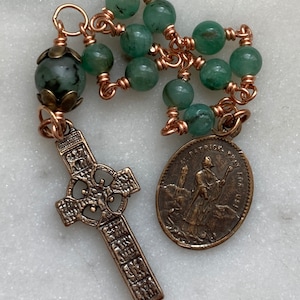 May include: A green stone rosary with a copper wire wrap and a Celtic cross charm. The rosary also features a round medal with the image of Saint Patrick and the text "Saint Patrick Pray For Us".