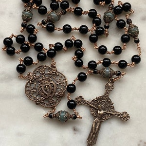 May include: A rosary featuring black beads strung on copper wire. The rosary includes a decorative pendant, and a crucifix. The beads are interspersed with gray beads. The metal components have an antique finish.