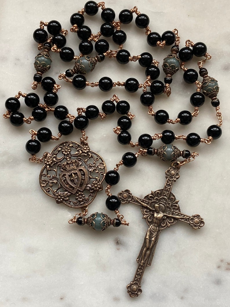 May include: A rosary featuring black beads strung on copper wire. The rosary includes a decorative pendant, and a crucifix. The beads are interspersed with gray beads. The metal components have an antique finish.