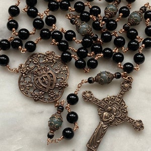 May include: A rosary featuring black beads and ornate bronze-colored metalwork. The design includes a decorative cross, a heart-shaped pendant, and detailed bead separators. The beads are strung on copper-colored wire.