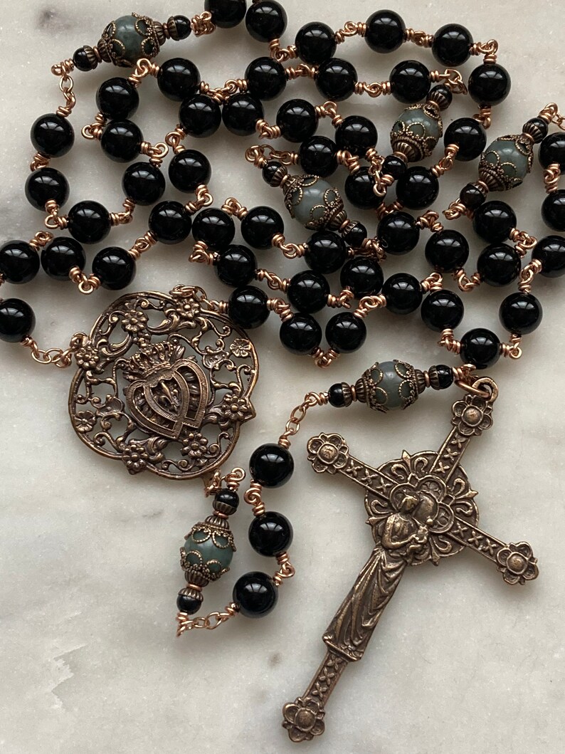 May include: A rosary featuring black beads and ornate bronze-colored metalwork. The design includes a decorative cross, a heart-shaped pendant, and detailed bead separators. The beads are strung on copper-colored wire.