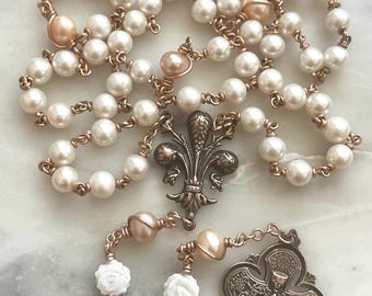 Puede incluir: Un rosario con perlas blancas y detalles en color melocotón. El rosario presenta una pieza central de bronce con forma de flor de lis y un colgante de cruz. Las cuentas están ensartadas en alambre color cobre.