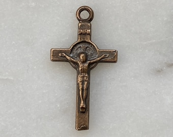 May include: A small, bronze-colored crucifix pendant with a detailed figure of Jesus Christ on the cross. The cross has a circular halo around the figure's head.