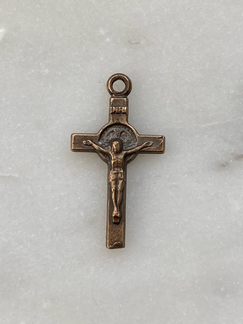 May include: A small, bronze-coloured crucifix pendant with a detailed figure of Jesus Christ on the cross. The cross has a circular halo around the figure's head.