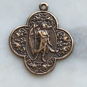 May include: A bronze-colored metal pendant featuring an angel with wings holding a sword. The pendant is in the shape of a four-leaf clover with intricate floral designs.
