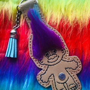 May include: A brown leather keychain with a purple and blue troll figure. The troll has a big smile and is wearing a purple wig. The keychain has a silver key ring and a blue tassel.