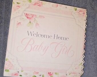May include: A pink and white baby girl welcome home sign with a lace border and floral design. The sign says "Welcome Home Baby Girl".