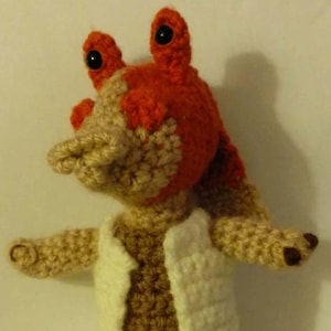 May include: A crocheted plush toy of a character from the Star Wars franchise. The toy is brown and tan with a red head and large black eyes. The toy is wearing a white crocheted robe.