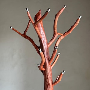 Puede incluir: Un perchero de madera artesanal con diseño de ramas de árbol. El perchero está teñido de un rico color marrón rojizo, con múltiples ramas que se extienden hacia afuera. Cada punta de rama tiene una pequeña bola plateada. Diseñado para abrigos y sombreros.