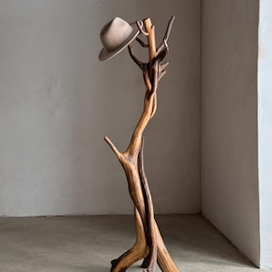 May include: A natural wood hat rack with a fedora hat. The rack is made from a twisted tree branch, with a light brown hat resting on top. The rack is a unique piece of home decor.