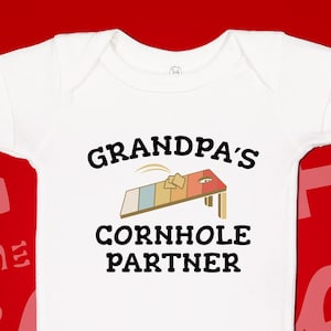 May include: White baby onesie with the text "GRANDPA'S CORNHOLE PARTNER" in black. The design includes a cornhole board graphic with beige, blue, and red colors. The onesie is short-sleeved.