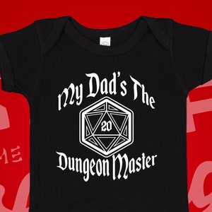 May include: Black baby onesie with white text that reads "My Dad's the Dungeon Master" and a white 20-sided die graphic.