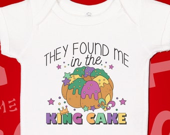 May include: White baby onesie with a colorful cartoon king cake and the text "They found me in the king cake".