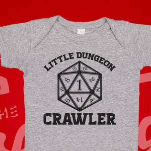 May include: A gray baby onesie with a black graphic of a twenty-sided die and the text "Little Dungeon Crawler".