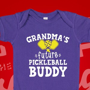 May include: Purple baby onesie with the text "Grandma's future pickleball buddy" and a yellow pickleball paddle graphic.