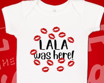 May include: White baby onesie with black text that reads "LALA was here!" surrounded by red lips.