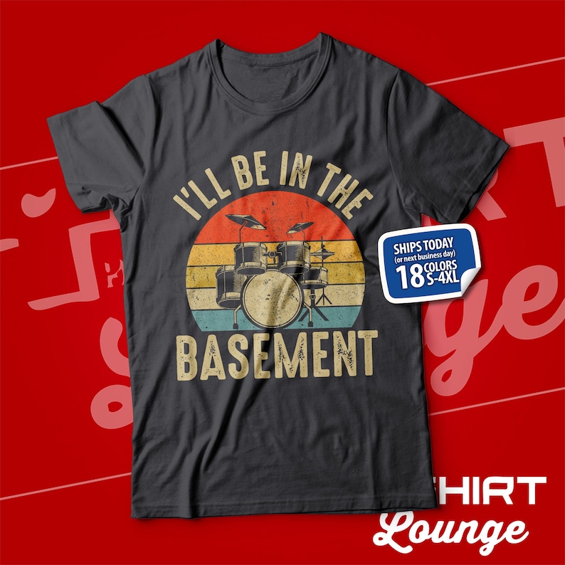 May include: A dark grey t-shirt featuring a vintage-style graphic of a drum kit in front of a sunset. The text "I'll be in the basement" is printed below the drum kit. The text "Ships Today (or next business day) 18 Colours S-4XL" is printed on a white sticker in the top right corner of the shirt.