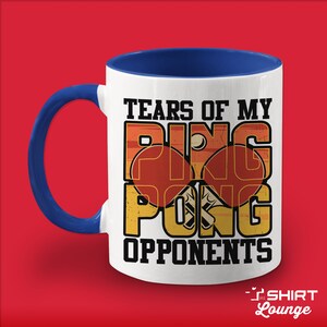 May include: White ceramic coffee mug with a blue rim and handle. The mug features a vintage-style graphic of two ping pong paddles and the text "Tears of my ping pong opponents".