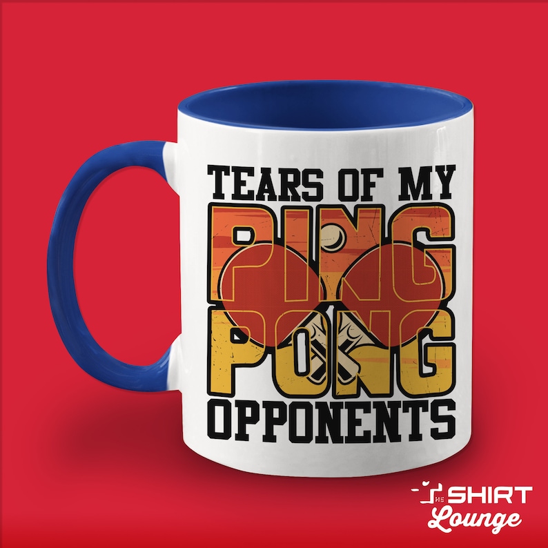 May include: White ceramic coffee mug with a blue rim and handle. The mug features a vintage-style graphic of two ping pong paddles and the text "Tears of my ping pong opponents".