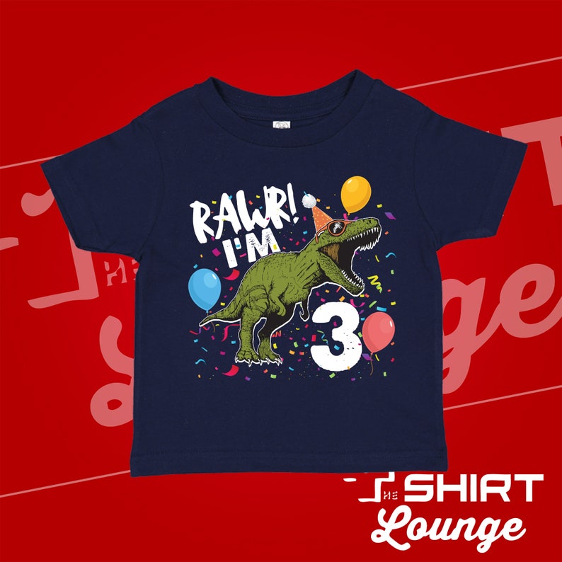 May include: A navy blue t-shirt with a green dinosaur wearing a party hat and sunglasses. The dinosaur is surrounded by confetti and balloons. The text "Rawr! I'm 3" is printed on the shirt.
