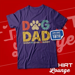 May include: Purple t-shirt with a retro-style graphic that says "Dog Dad" with a paw print in the "O". The graphic is in shades of red, orange, yellow, green, and blue. Below the graphic, it says "Scout Luna Buddy" with a heart between each name.