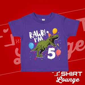 May include: Purple short-sleeved t-shirt with a green dinosaur wearing sunglasses and a party hat. The dinosaur is saying "Rawr! I'm 5" with confetti and balloons around it.