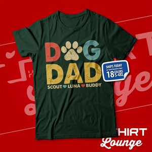 May include: Green t-shirt with a retro-style graphic that says "Dog Dad" with a paw print in the "O". The graphic is in red, yellow, blue, and green. Below the graphic, it says "Scout Luna Buddy" with a heart between each name.