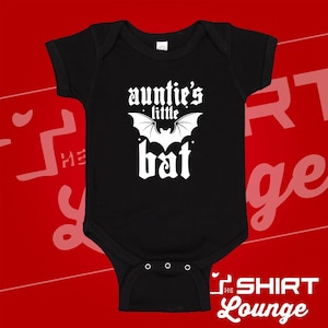 May include: Black baby bodysuit with the text "auntie's little bat" in white, along with a bat graphic. The bodysuit has short sleeves and three snap closures at the bottom. The Shirt Lounge logo is visible.