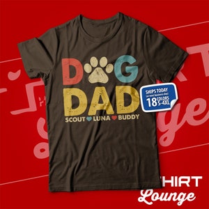 May include: A brown t-shirt with the text "Dog Dad" in a retro style with a paw print in the middle of the "O". The text "Scout Luna Buddy" is below the main text with a heart between each name.