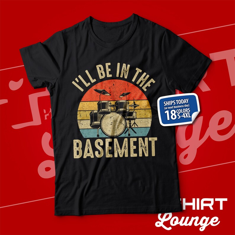 May include: Black t-shirt featuring a vintage-style graphic of a drum kit in front of a sunset. The text "I'll be in the basement" is printed below the drum kit.