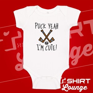 May include: White baby onesie with the text "Puck Yeah I'm Cute!" and a graphic of crossed hockey sticks and a puck. The onesie is short-sleeved and has a snap closure at the bottom.