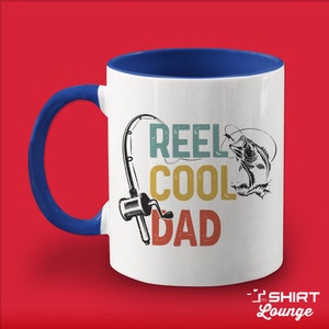 May include: White ceramic coffee mug with a blue handle. The mug has a vintage-style graphic of a fishing rod, reel, and fish with the text "REEL COOL DAD".