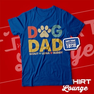 May include: Blue t-shirt with a vintage-style graphic that says "Dog Dad" with a paw print in the "O". The graphic is in red, yellow, blue, and green. Below the graphic, it says "Scout Luna Buddy" with a heart between each name.